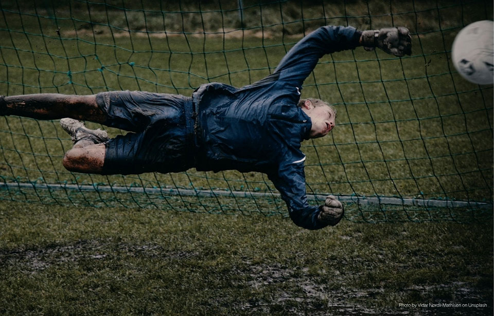 A muddy football goalkeeper in a blue kit diving mid-air to save a ball next to the goal net on a wet, grass pitch.